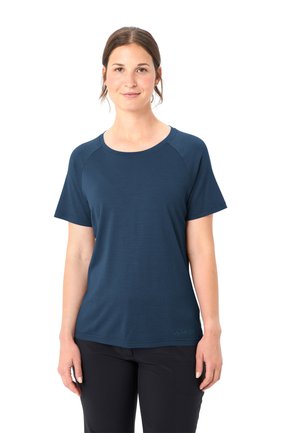 Frau mit braunen Haaren im Dutt, die ein schlichtes dunkelblaues Kurzarmshirt und schwarze Hosen trägt, steht vor einem weißen Hintergrund.