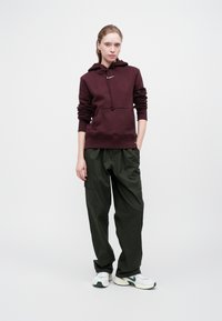 Sweatshirt capuz bordeaux com bolso frontal, combinado com calças cargo verdes-azeitona de corte largo. O modelo usa ténis brancos com detalhes verdes.