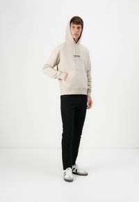 Beige Kapuzenpullover mit einer Fronttasche und Kordelzug-Kapuze, kombiniert mit schwarzen Jeans und weißen Sneakers mit grauen Akzenten.