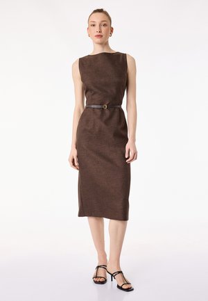 Kobieta ubrana w brązową sukienkę midi bez rękawów z cienkim czarnym paskiem i czarne, wiązane sandały na obcasie, stojąca na białym tle.