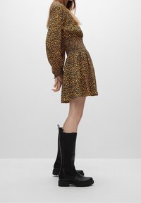 Zwarte laarzen tot de knie met een matte afwerking, dikke zool en een rits aan de zijkant. De jurk heeft een aansluitend gerimpeld lijfje en een geel en zwart patroon.