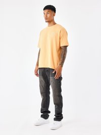 Kortlopend oranje t-shirt met geborduurd ontwerp, gecombineerd met zwarte jeans met een vervaagde textuur en voorzakken, afgemaakt met witte sneakers.