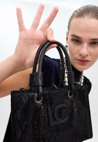 Un mannequin pose avec un sac à main en sequins noirs arborant le logo "LC", illustrant un geste de la main tout en portant des accessoires audacieux.