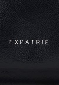 Surface en cuir noir texturé avec le mot « EXPATRIÉ » embossé en lettres capitales blanches au centre.