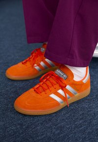 Ljusa orange Adidas-sneakers med vita ränder och gummisulor, bärs med vita strumpor och lila byxor på en blå matta.