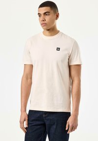 Tricou din bumbac bej deschis, cu mâneci scurte și guler rotund. Prezintă un mic logo pătrat negru pe pieptul stâng. Design simplu.
