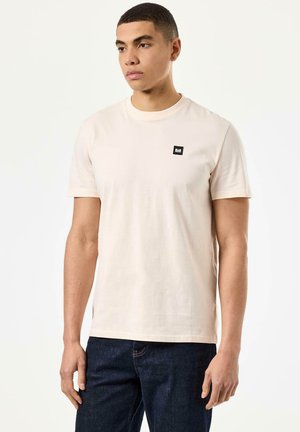 Lys beige bomull t-skjorte med korte ermer og rund hals. Har en liten svart firkantet logo på venstre bryst. Enkelt design.