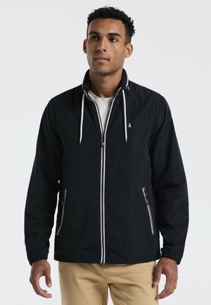 Mann trägt eine schwarze Jacke mit Reißverschluss und weißen Kordeln und beige Hose, steht vor einem schlichten hellen Hintergrund.