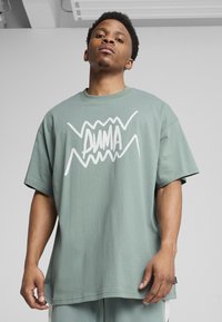 Grünes übergroßes T-Shirt aus Baumwolle mit einem weißen Grafiklogo in der Mitte. Kurze Ärmel und runder Ausschnitt. Weiche Textur.