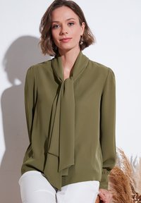 Olive green blouse in lightweight fabric, featuring a tied collar detail and long sleeves. Paired with white pants, smooth texture.