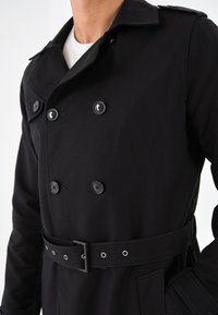 Svart trenchcoat med dubbelknäppning, bälte med metalldolk, och sidofickor. Slät tygstruktur, skräddarsydd passform.