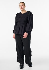 Naine kannab musta smockiga pikade varrukatega pluusi, lahtisi musti pükse ja musti terava nina ning pandlatega kingi, seistes valge tausta ees.