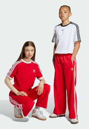 Rotes Sportoutfit bestehend aus gestreiften Ärmeln und Hosen. Ein Modell kniet und trägt ein rotes Oberteil mit passender Hose; das andere steht in Weiß und Schwarz.