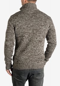 Brown knitted sweater with a high collar, ribbed cuffs and hem, featuring a textured pattern with lighter flecks throughout. Dark jeans visible.