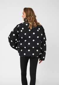 Svart boblejakke med store hvite prikkemerker, med høy krage og polstret tekstur. Modellen har på seg svarte leggings.