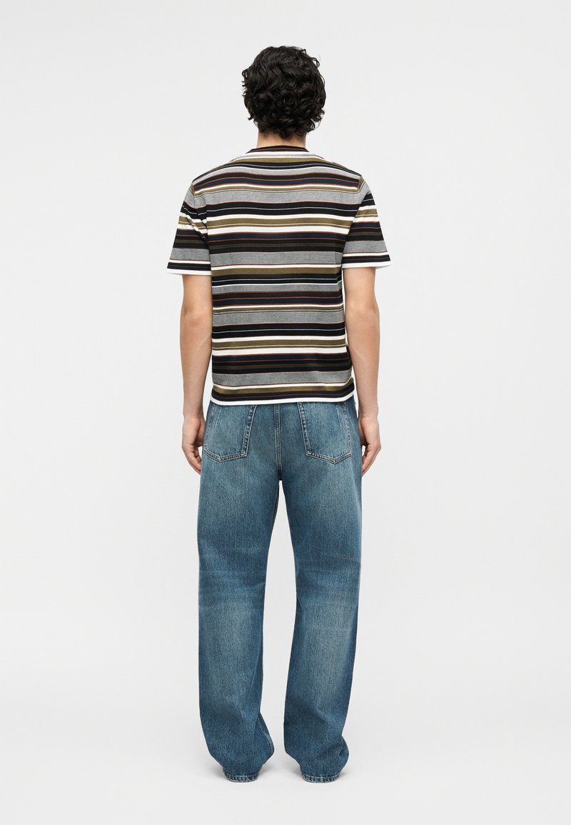 Man standing facing away wearing a short-sleeve horizontal striped shirt and loose blue jeans against a plain white background.