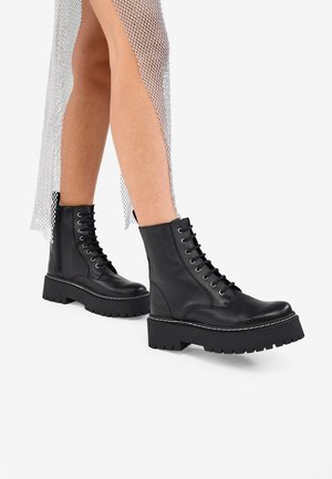 Botas de piel negra hasta el tobillo con suelas gruesas, diseño de cordones y detalles de costura blanca. La parte superior de tela texturizada tiene un patrón de red de pescar.