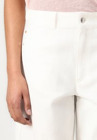 Pantaloni in denim bianco con vita aderente, dotati di chiusura con bottone visibile e cucitura laterale. Il tessuto ha una consistenza liscia e una leggera elasticità.