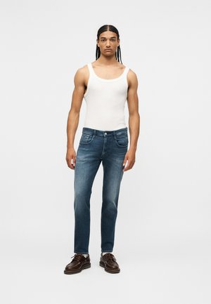 Young man with braided hair wearing a white tank top, blue jeans, white socks, and dark brown leather shoes, standing against a plain background.