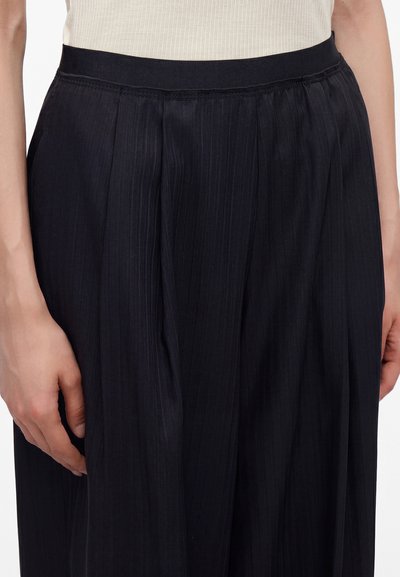 Gros plan d'une personne portant un pantalon plissé noir taille haute avec un top crème côtelé rentré à l'intérieur et une main reposant sur le côté.