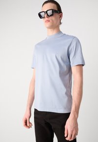 Lys blå kortermet T-skjorte laget av glatt stoff, brukt med store svarte solbriller. Enkel rund hals og minimalt design.