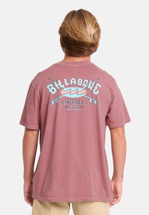 Persona con capelli biondi corti che indossa una maglietta rossa sbiadita Billabong Australia con logo e testo sul retro, abbinata a pantaloni beige.