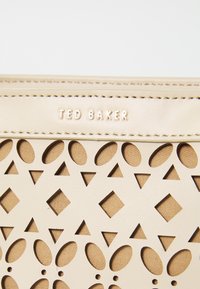 Beige läppärkkä käsilaukku, jossa on geometrisiä leikkeitä, nahkatekstuuri ja kultaiset "TED BAKER" -logot. Liukas pinta ja vaalea kangaspohja.