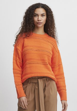 Pull en laine orange avec des rayures horizontales et des détails ajourés. Présente des poignets côtelés et un col rond. Associé à un pantalon marron.