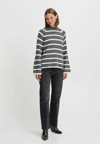 Pull en tricot rayé gris et blanc avec un col haut et des manches évasées, porté avec un jean noir et des bottes à bout pointu.