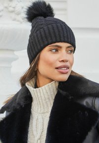 Beanie lavorato a maglia nera con un morbido pom-pom in cima. Il berretto ha un design a coste testurizzato, abbinato a un maglione a collo alto in maglia leggera.