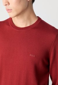Pull en tricot rouge avec un col rond et un logo "BOSS" brodé sur la poitrine. Texture lisse avec une coupe ajustée.