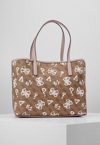 Sac cabas marron avec des poignées roses, décoré de points d'interrogation blancs, de triangles, de chiffres et de logos « GS » entrelacés, présenté sur une surface blanche.