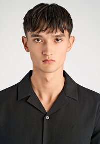 Maglietta nera a maniche corte con colletto a destra, tessuto leggero e sottile texture. Presenta un piccolo logo ricamato sul petto.