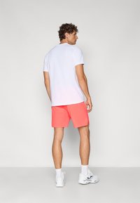 T-shirt branca de manga curta combinada com calções corais brilhantes. Ténis brancos com detalhes pretos. Vista traseira, em pé sobre fundo simples.