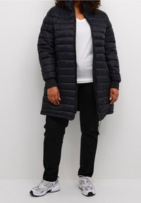Giacca lunga trapuntata nera con colletto rialzato, chiusura a zip davanti e polsini a costine, abbinata a pantaloni neri e sneakers bianche.