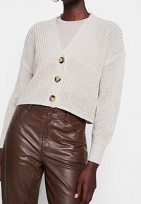 Beige stickad cardigan med V-ringning, prydd med tre stora knappar och ribbstickade ärmslut, som är ihop med bruna glansiga läderbyxor.