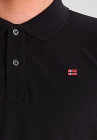 Svart polo tröja i texturerad tyg, med krage och tvåknapps knäppning. Utmärkande broderad detalj med röd och vit flagga.