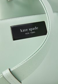 Borsa a mano in pelle verde menta con una targhetta nera con il logo "kate spade NEW YORK." Texture liscia con cuciture e manici a contrasto.