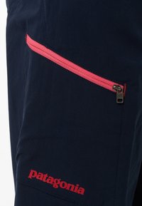 Tmavě modré kalhoty s diagonálním růžovým zipem na kapse a červeným logem "Patagonia" na lemu. Vyrobené z lehké tkaniny.