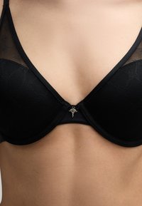 Zwarte beha met een bovenste laag van gaas, onderrugsteun, kantdetails en een decoratieve charm in het midden. Over het geheel een gladde textuur.