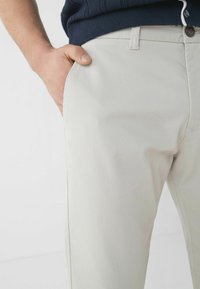 Pantaloni chino beige chiaro con tessuto liscio e leggermente strutturato. Presentano tasche laterali e chiusura con bottone, abbinati a un maglione blu navy alla vita.