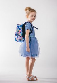 Zaino con motivo floreale luminoso e multicolore, spallacci imbottiti e tasche con zip. Bambina in vestito blu e sandali argento si trova accanto ad esso.