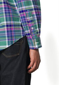 Green and purple plaid long-sleeve shirt with button cuffs, paired with dark denim. Features varied thickness stripes and a pocket.
