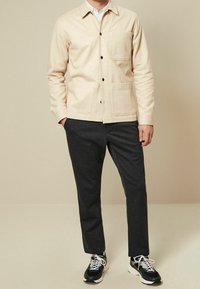 Giacca camicia in cotone beige con chiusura a bottoni, due tasche frontali e colletto. Abbinata a pantaloni grigi scuri e sneaker nere.