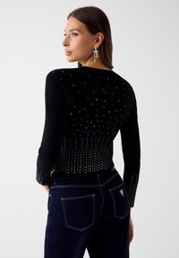 Czarny dziany sweter z metalicznymi ćwiekami, o dopasowanym kroju i długich rękawach. Połączony z ciemnymi dżinsami, z widocznymi detalami przeszyć.