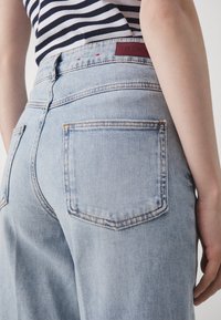 Person som bär ljusblå denimjeans och en svart- och vitrandig skjorta, sedd från midjan ner och bakifrån.