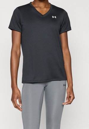 Personne portant un t-shirt noir à col en V et manches courtes ainsi que des leggings gris clair avec de petits logos Under Armour sur le t-shirt et les leggings.