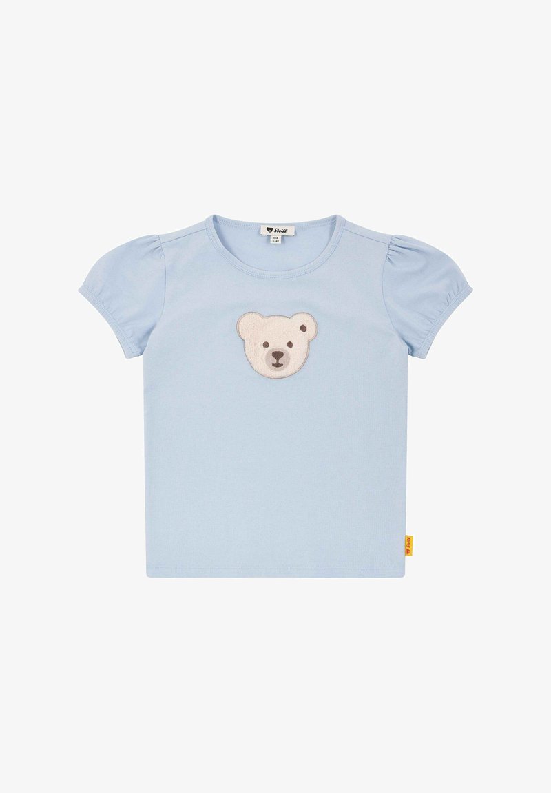 Jasnoniebieska koszulka z krótkimi bufkami, z beżowym i brązowym aplikacją w kształcie niedźwiedzia na środku oraz żółtą metką z boku.