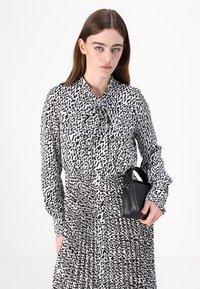 Schwarz-weiß gemustertes Langarmkleid mit gebundenem Kragen, das einen plissierten Rock und eine kleine schwarze Handtasche enthält, die in einer Hand gehalten wird.