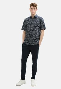 Chemise navy à manches courtes avec un motif de feuilles blanches, assortie à un pantalon foncé et des baskets blanches. La chemise a un col et une fermeture éclair sur le devant.